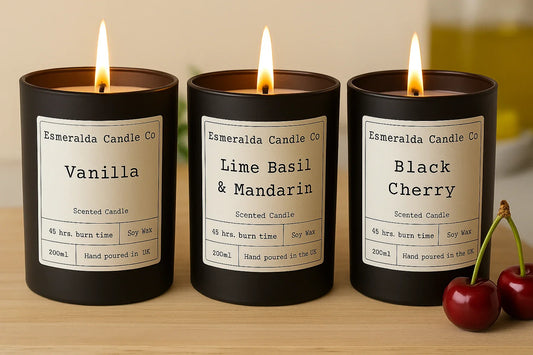 Three lit luxury scented candles from Esmeralda Candle Co in black jars, labeled Vanilla, Lime Basil & Mandarin, and Black Cherry, hand-poured soy wax with 45-hour burn time.