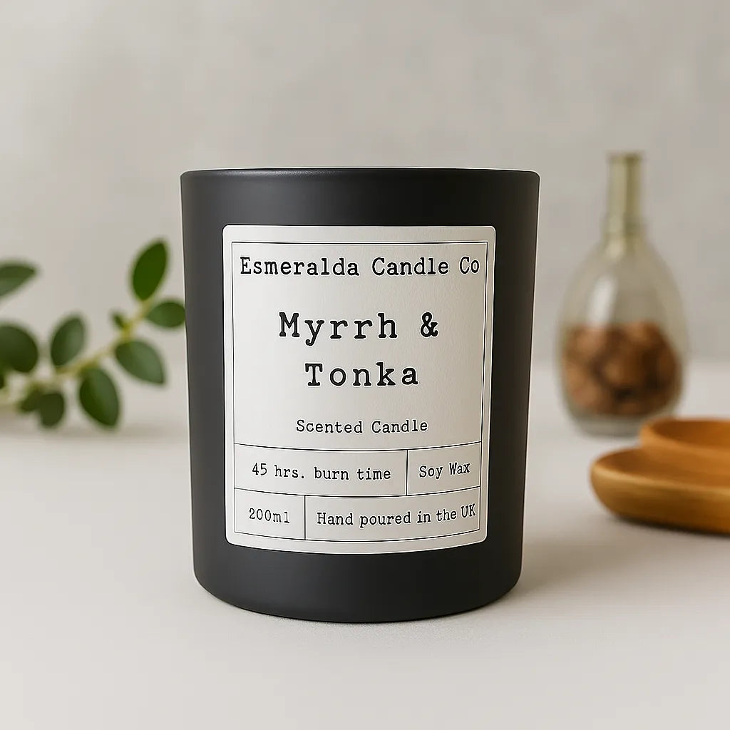 Esmeralda Candle Co Myrrh & Tonka scented candle in a matte black jar with a minimalist white label displaying '45 hrs. burn time', 'Soy Wax', '200ml', and 'Hand poured in the UK', styled on a neutral surface with eucalyptus, a wooden tray, and a glass jar in the background