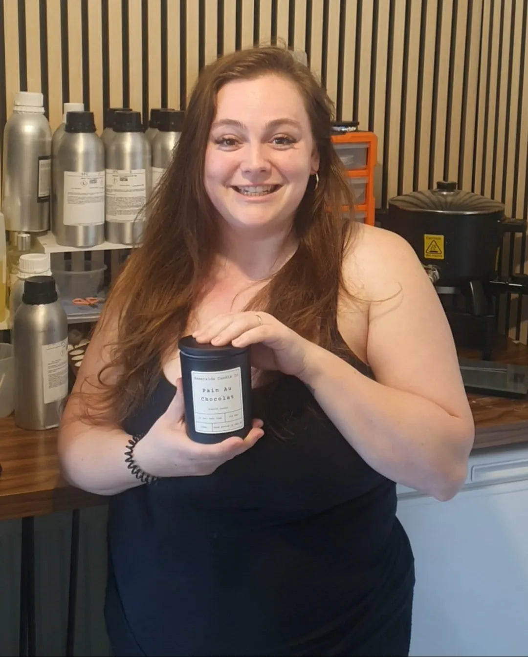 Smiling woman holding a 'Pain Au Chocolat' scented candle from Esmeralda Candle Co in a candle-making studio, surrounded by fragrance oil bottles and equipment.