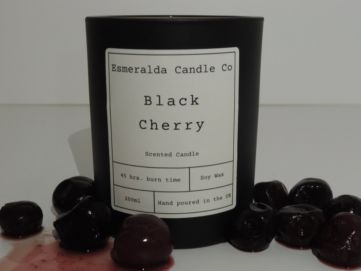 A black cherry scented candle in a matt black glass jar on a white shelf with black cherries spilled out around it.