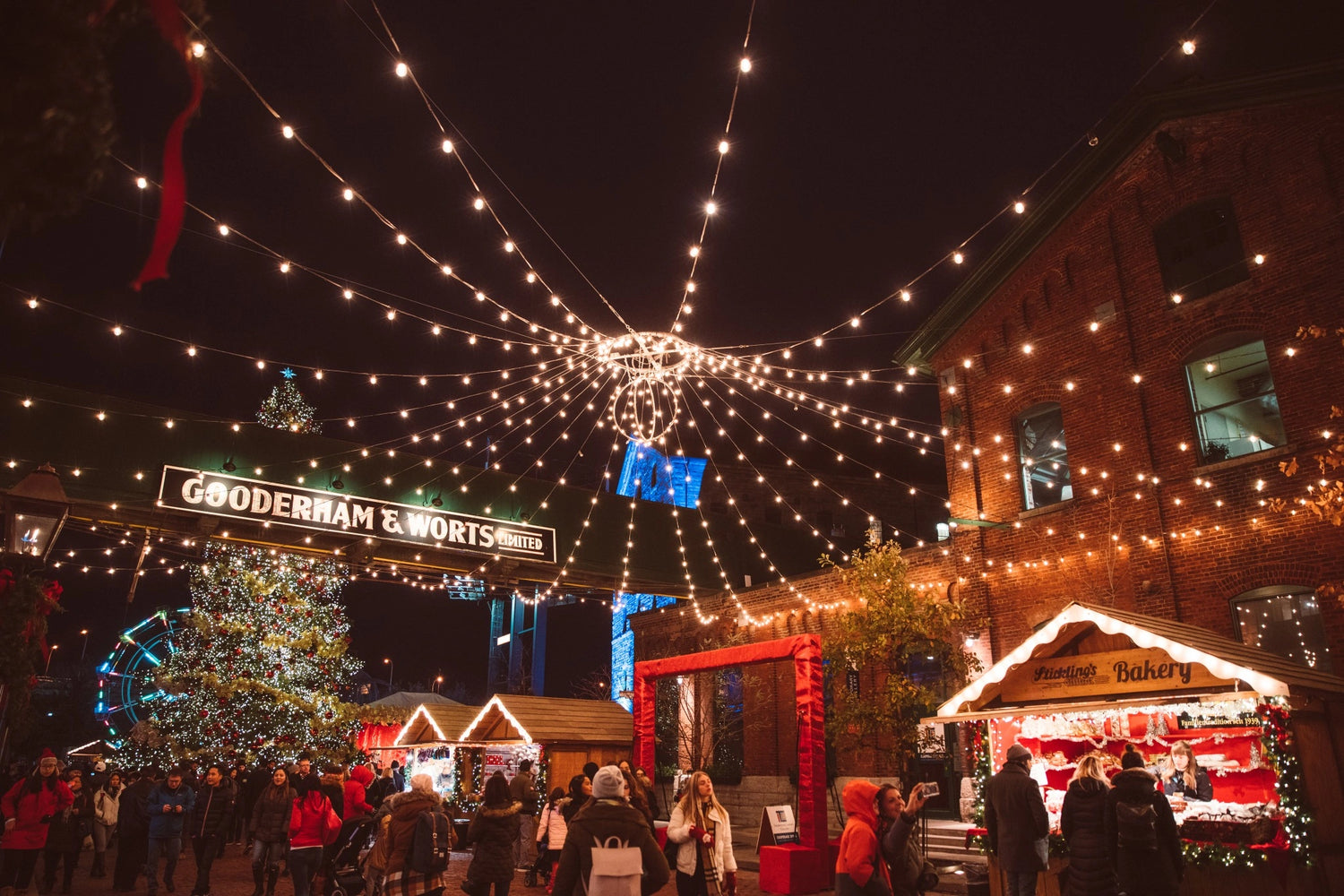 Crowds enjoying a festive Christmas market at night with string lights, decorated stalls, and a large Christmas tree