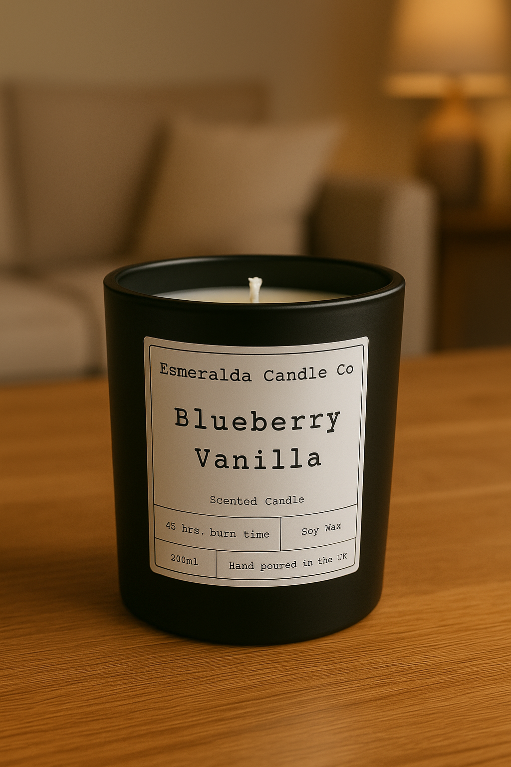 A matte black Esmeralda Candle Co. “Blueberry Vanilla” scented candle sits on a polished wooden table in a cozy living room, with warm lighting and blurred neutral-toned decor in the background.