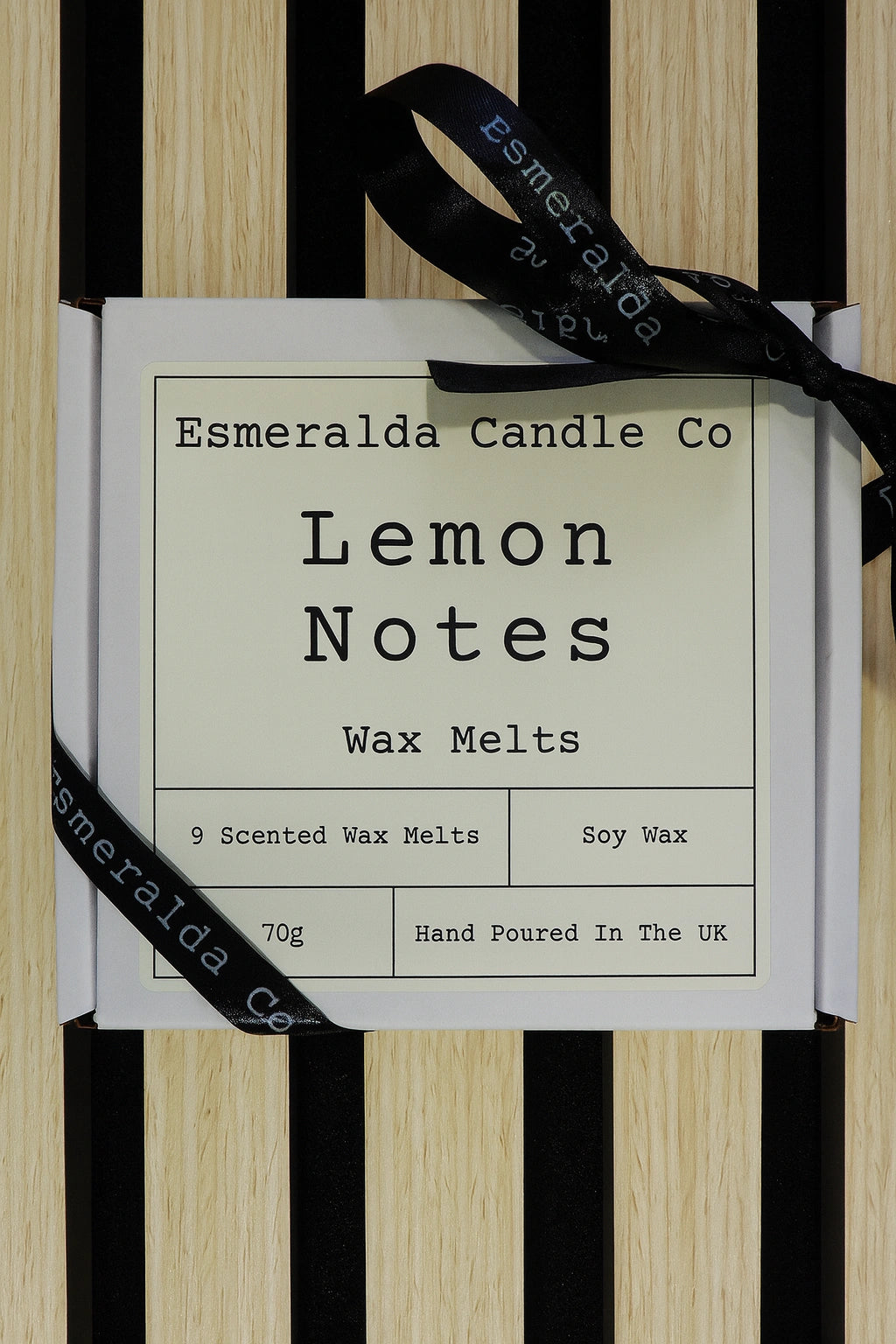 A square white box labelled 'Esmeralda Candle Co – Lemon Notes Wax Melts' tied with a black ribbon, displayed on a striped wood background.