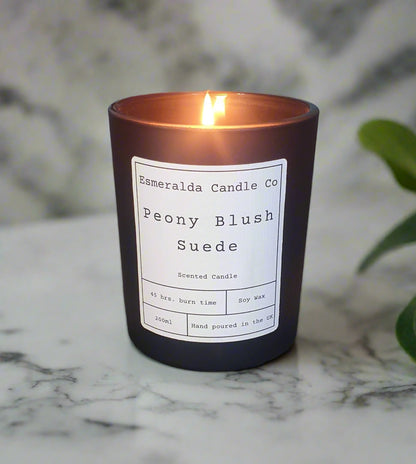 Esmeralda Candle Co. Peony Blush Suede scented candle lit on a dining table with orchid plant in the background. Hand-poured soy wax candle, 200ml, 45-hour burn time, made in the UK.