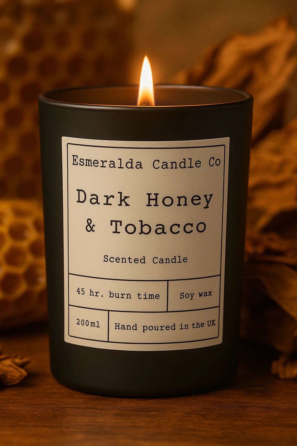 Black candle labeled 'Esmeralda Candle Co Dark Honey & Tobacco' with a flame on a wooden surface.