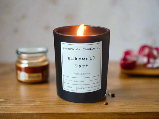 A lit black jar candle with a white label reading 'Esmeralda Candle Co – Bakewell Tart.' The candle sits on a wooden surface with a warm glow, with jars and red decor blurred in the background.