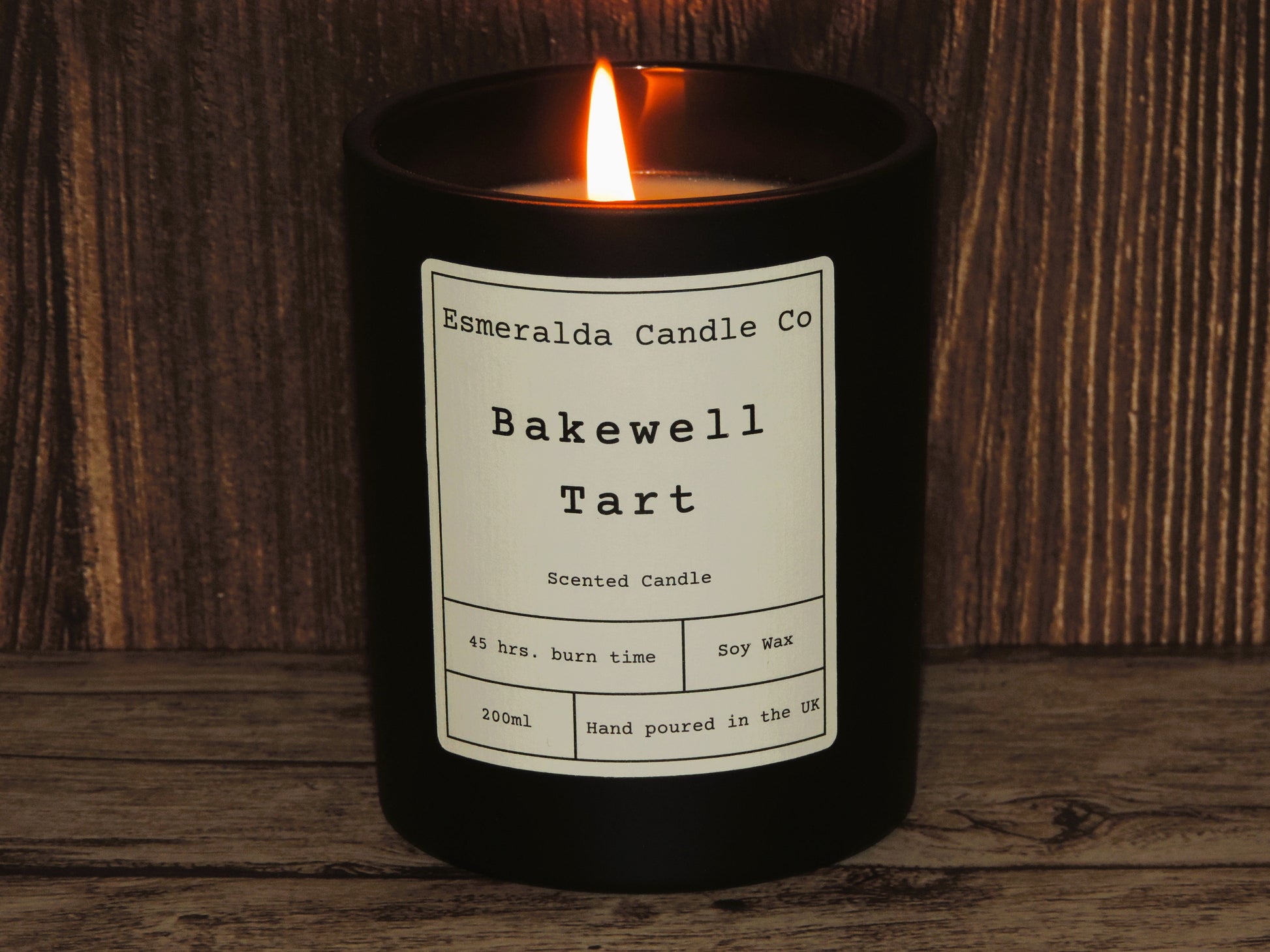 A lit bakewell tart scented black glass candle, on a wooden shelf