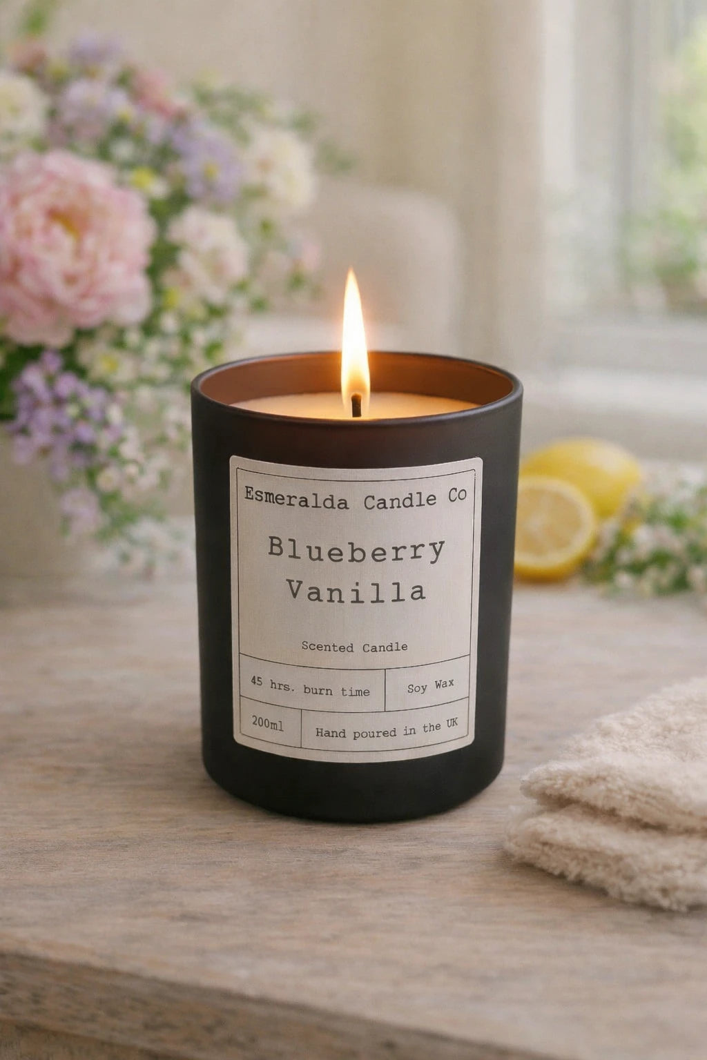 Esmeralda Candle Co Blueberry Vanilla candle on a table with flowers and lemons in the background