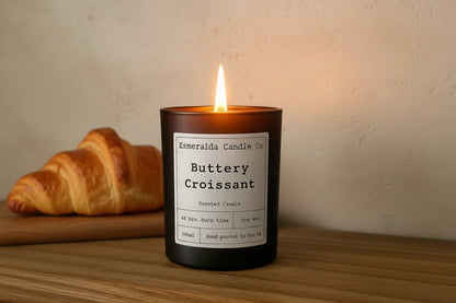 Scented candle labeled 'Buttery Croissant' by Esmeralda Candle Co on a wooden surface.