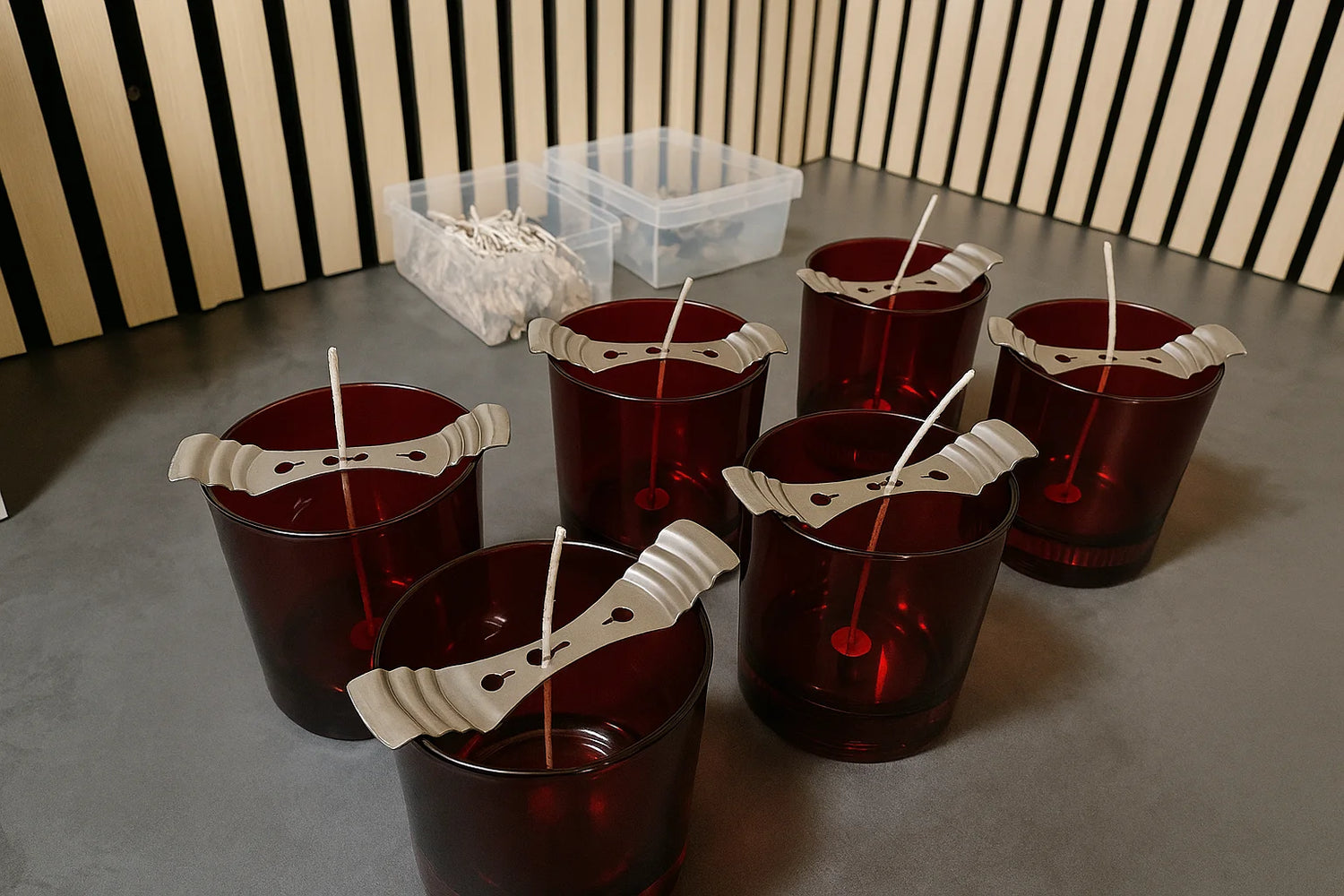 Six red glass jars arranged on a counter with wick holders keeping candle wicks upright, with containers of extra wicks and tabs in the background.