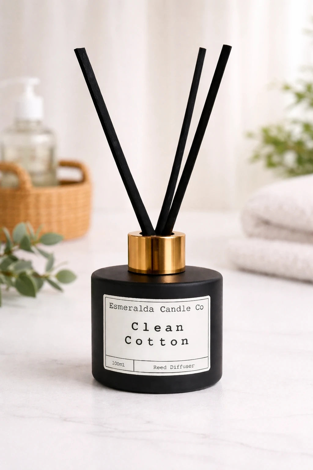 Esmeralda Candle Co Clean Cotton reed diffuser in a matte black bottle with gold collar and black reeds, 100ml home fragrance product on a light blue background.