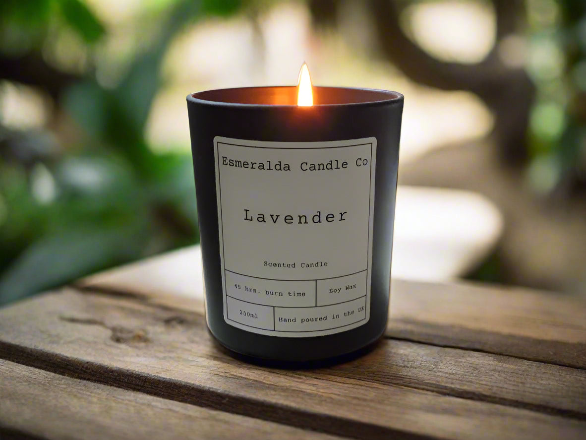 Lit Lavender scented soy wax candle by Esmeralda Candle Co, hand poured in the UK, displayed on a wooden surface with a natural green garden background for a calming outdoor ambiance