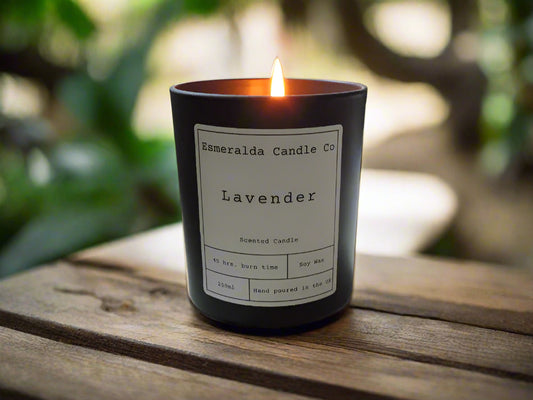 Lit Lavender scented soy wax candle by Esmeralda Candle Co, hand poured in the UK, displayed on a wooden surface with a natural green garden background for a calming outdoor ambiance