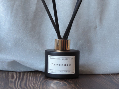 Lavender reed diffuser by Esmeralda Candle Co, 100ml matte black bottle with gold collar and black reeds, displayed on dark wood with silver fabric backdrop for a calming, elegant home scent.