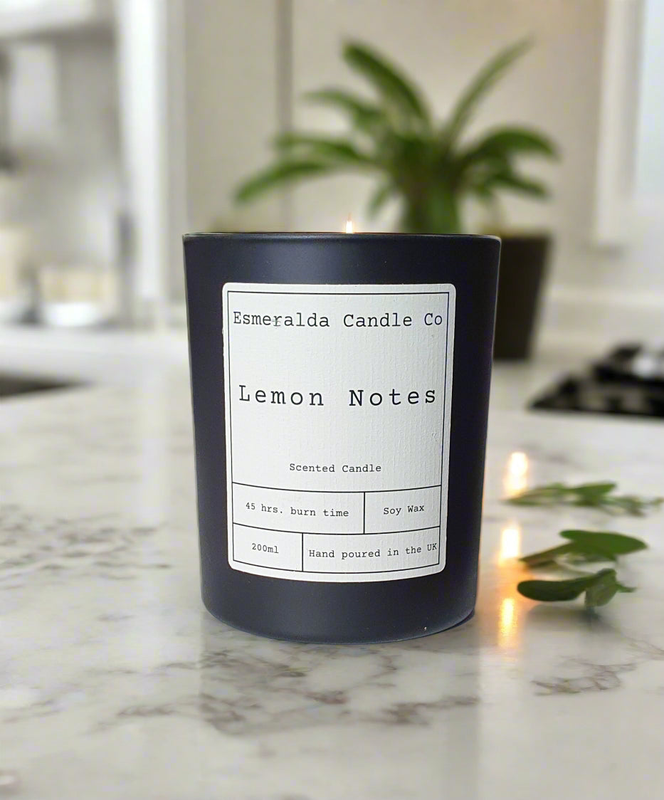 Black candle labelled 'Lemon Notes' on a worktop with green leaf's scattered and a plant in the background. candle is made by Esmeralda candle co