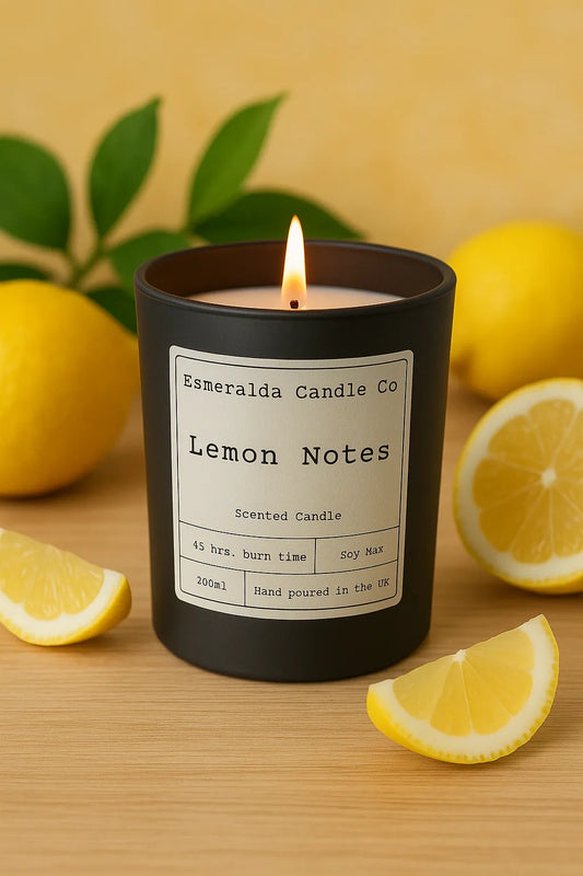 A lit Esmeralda Candle Co. “Lemon Notes” scented candle in a matte black glass jar, placed on a wooden surface surrounded by fresh lemons and green leaves, with a warm yellow backdrop.