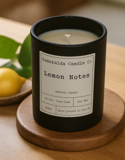 Black candle labelled 'Lemon Notes' on a worktop with green leaf's scattered and a plant in the background. candle is made by Esmeralda candle co