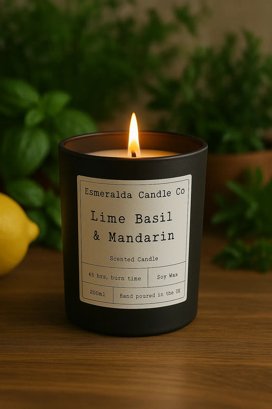 A lit Esmeralda Candle Co. “Lime Basil & Mandarin” scented candle in a matte black glass jar, placed on a wooden surface with fresh basil leaves, greenery, and a lemon in the blurred background.