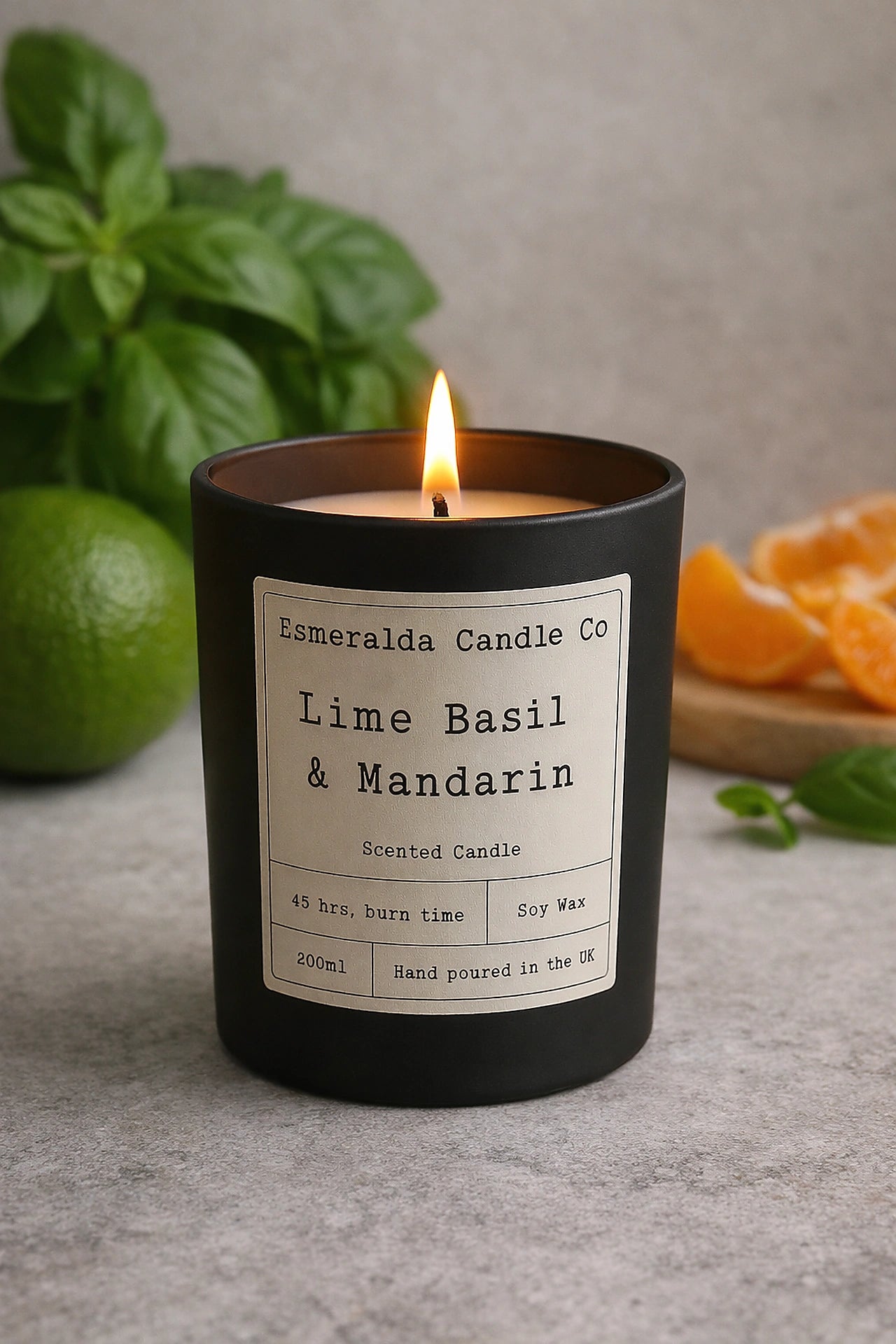 A lit Esmeralda Candle Co. “Lime Basil & Mandarin” scented candle in a matte black glass jar, placed on a wooden surface with fresh basil leaves, greenery, and a lemon in the blurred background.