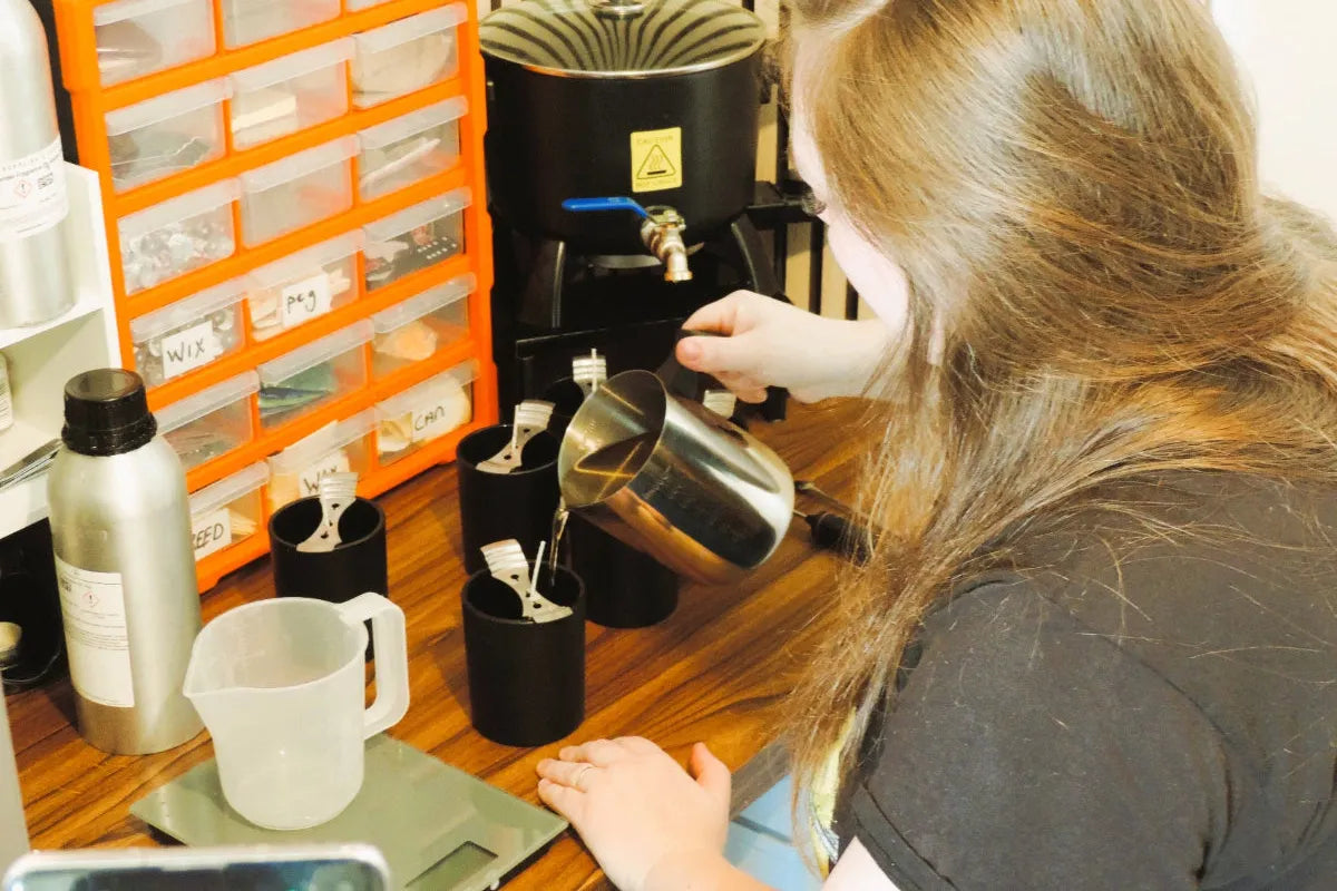 person hand-pouring melted wax into black candle jars on a wooden workbench, with tools, containers, and labelled storage drawers in the background.