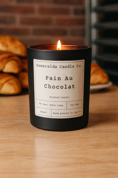 Candle labeled 'Pain Au Chocolat' from Esmeralda Candle Co on a wooden surface with bread in the background.