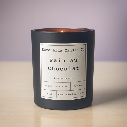 Pain Au Chocolat scented soy candle by Esmeralda Candle Co, lit inside a matte black glass jar with a clean white label, placed on a light wooden surface against a soft lavender background.