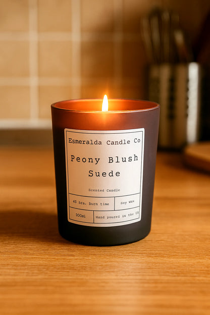 A lit Esmeralda Candle Co. “Peony Blush Suede” scented candle in a matte dark purple glass, placed on a wooden kitchen counter with blurred utensils and tiled backsplash in the background.