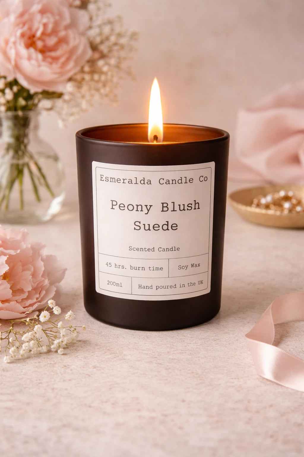 Scented candle labeled 'Peony Blush Suede' by Esmeralda Candle Co on a light surface with floral decorations.
