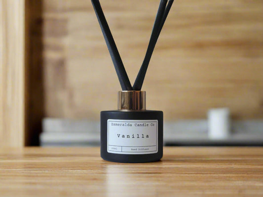 Vanilla reed diffuser by Esmeralda Candle Co, 100ml matte black bottle with gold collar and black reeds, styled against soft silver fabric for a cozy and elegant home scent