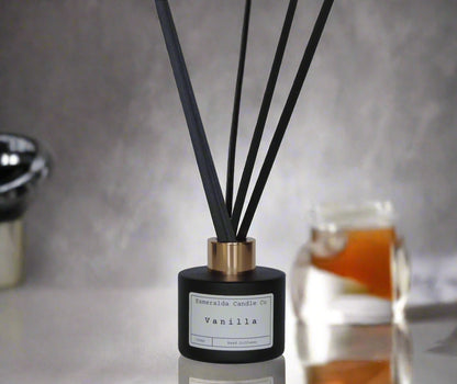 Vanilla reed diffuser by Esmeralda Candle Co, 100ml matte black bottle with gold collar and black reeds, styled on reflective surface with warm background accents for a cozy home ambiance.