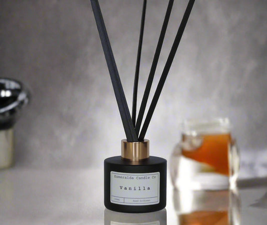 Vanilla reed diffuser by Esmeralda Candle Co, 100ml matte black bottle with gold collar and black reeds, styled on reflective surface with warm background accents for a cozy home ambiance.