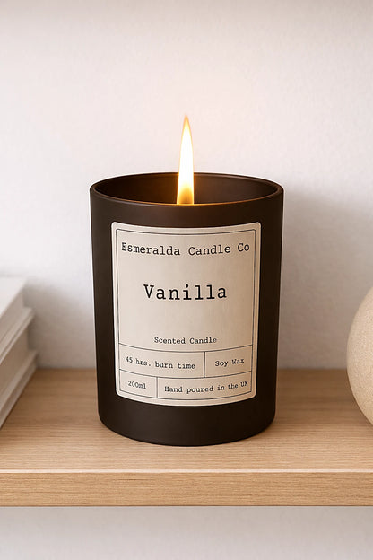 A lit Vanilla scented candle from Esmeralda Candle Co in a black glass jar with a white label, showing 45 hrs. burn time, Soy Wax, 200ml, and Hand poured in the UK